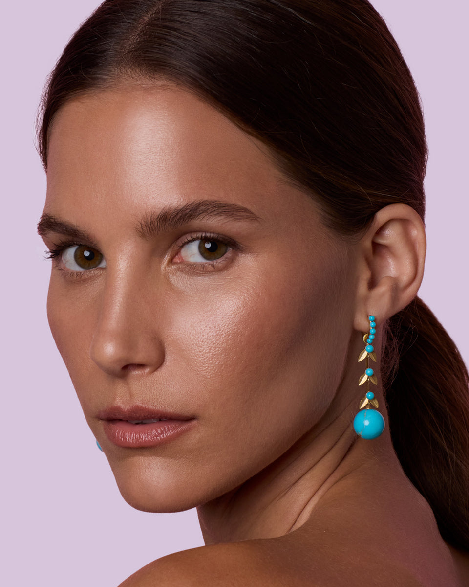 A close-up of a woman with dark hair pulled back, wearing a single dangling earring with turquoise blue beads and gold-toned triangular accents against a light lavender background.
