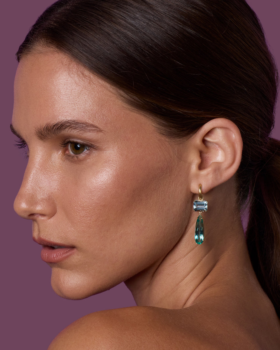 A close-up side profile of a woman wearing a gold drop earring with a rectangular light blue gemstone and a teardrop-shaped blue-green gemstone pendant, set against a solid mauve background.