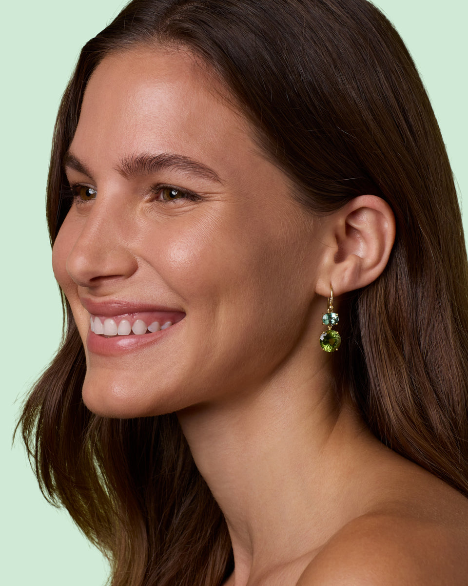 A close-up side profile of a smiling woman with long brown hair wearing a small gold dangle earring with green gemstones, set against a solid pale mint green background.