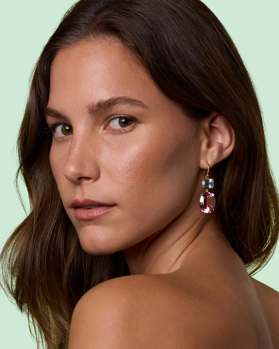 A close-up portrait of a woman with brown hair wearing a dangling earring with a silver-toned top and a pink teardrop-shaped gemstone, set against a pale mint green background.
