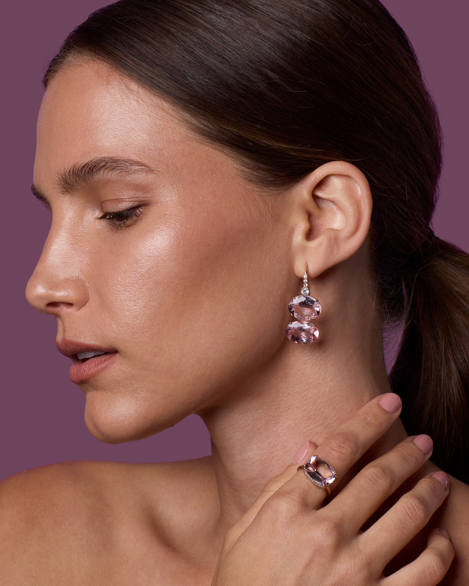 A close-up side profile of a woman with dark hair in a ponytail, wearing a dangling pink gemstone earring and a matching pink gemstone ring on her finger, against a solid mauve-purple background.