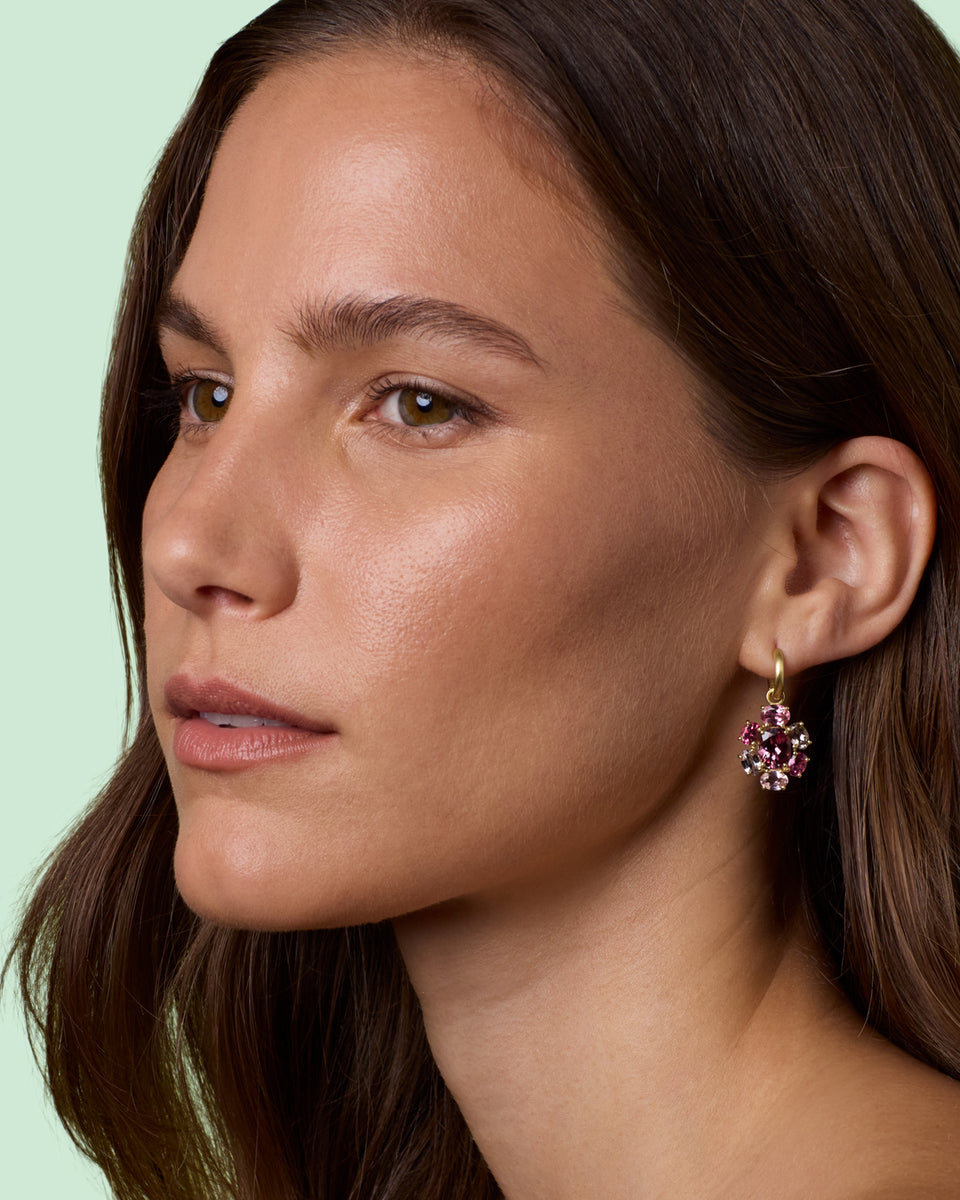 A close-up side profile of a woman with dark brown hair wearing a small gold dangle earring with pink gemstones, set against a solid pale mint green background.