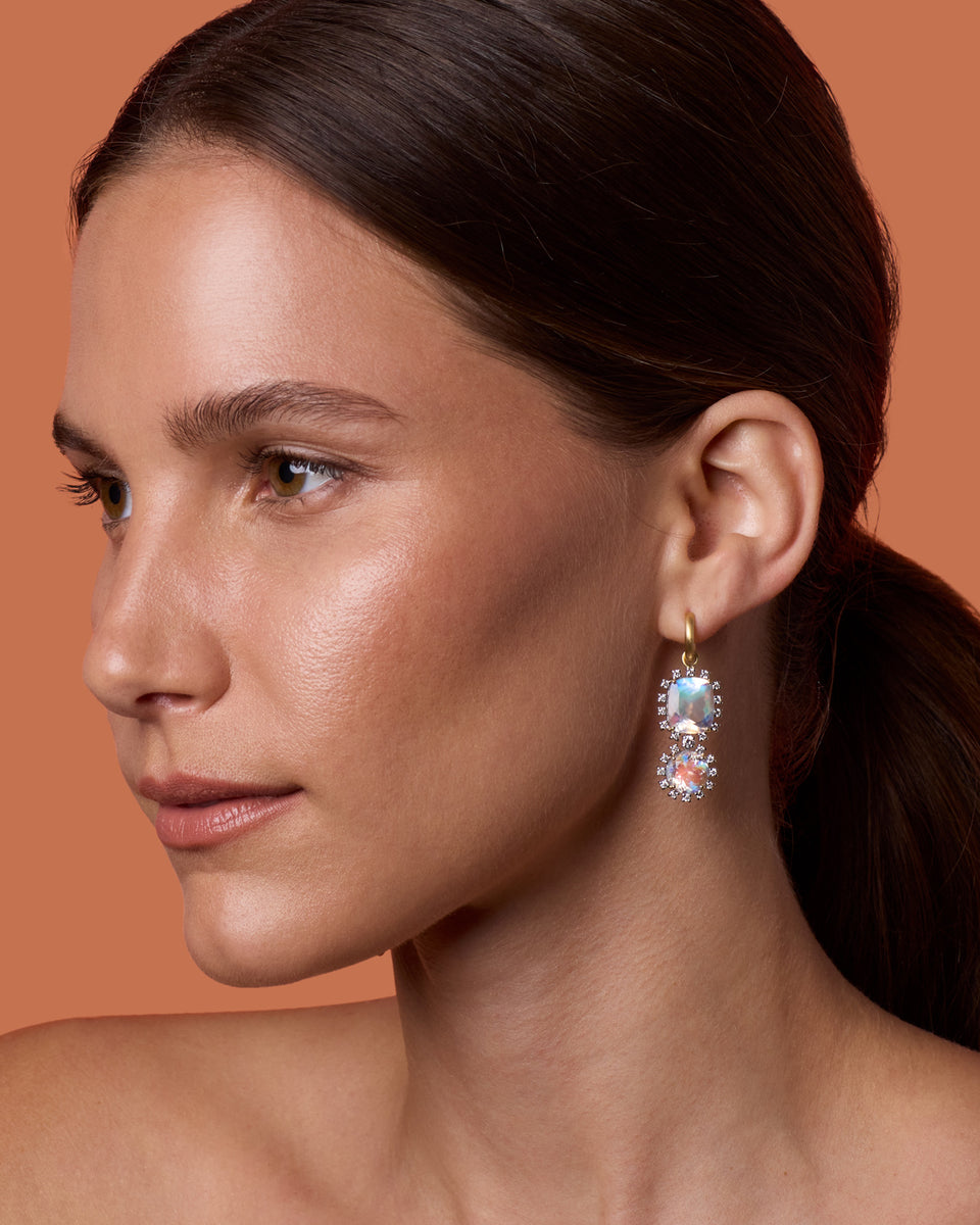 A close-up side profile of a woman with dark hair tied back, wearing a dangling earring with iridescent blue and pink stones in a gold setting, against a solid warm peach background.