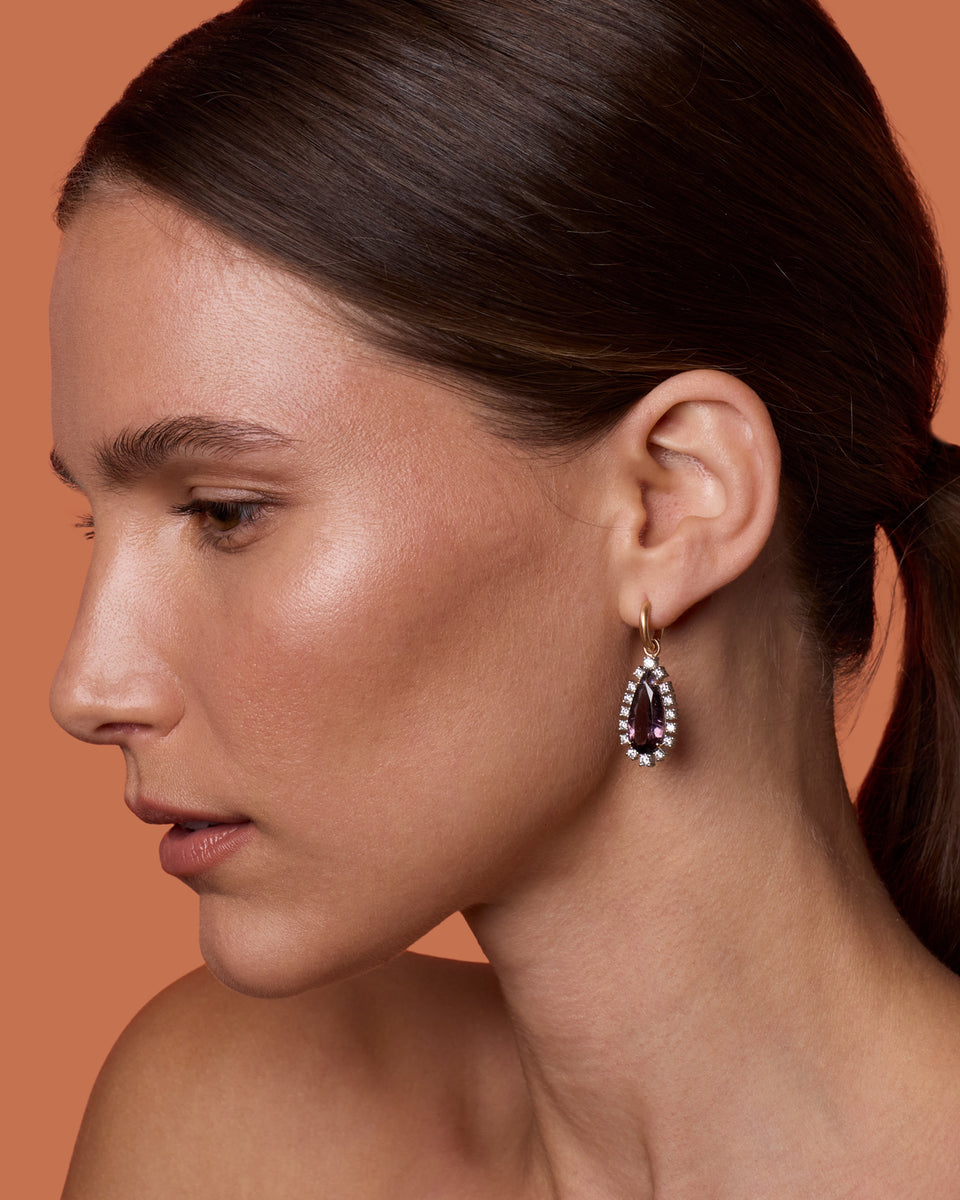 A close-up side profile of a woman with dark hair tied back, wearing a teardrop-shaped dangling earring with a dark purple center stone and a border of small clear stones, set against a solid warm peach background.