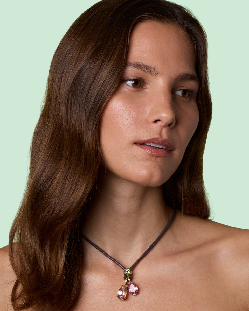 A close-up of a woman with long brown hair wearing a delicate dark cord necklace with a small gold-toned connector and two dangling pink gemstones, set against a solid pale mint green background.