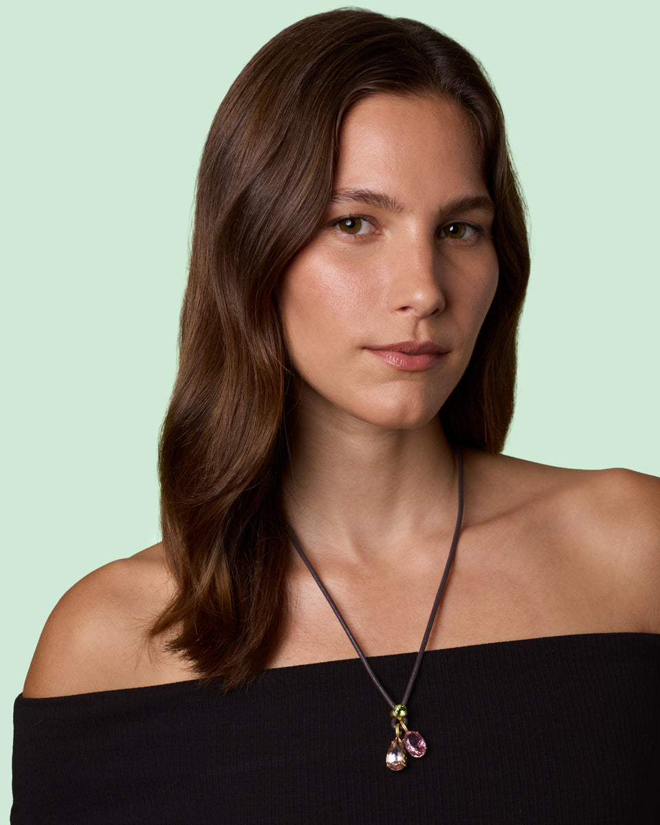 A woman with long brown hair wearing a black off-the-shoulder top and a delicate necklace with a dark cord and small pink pendants, set against a solid pale mint green background.