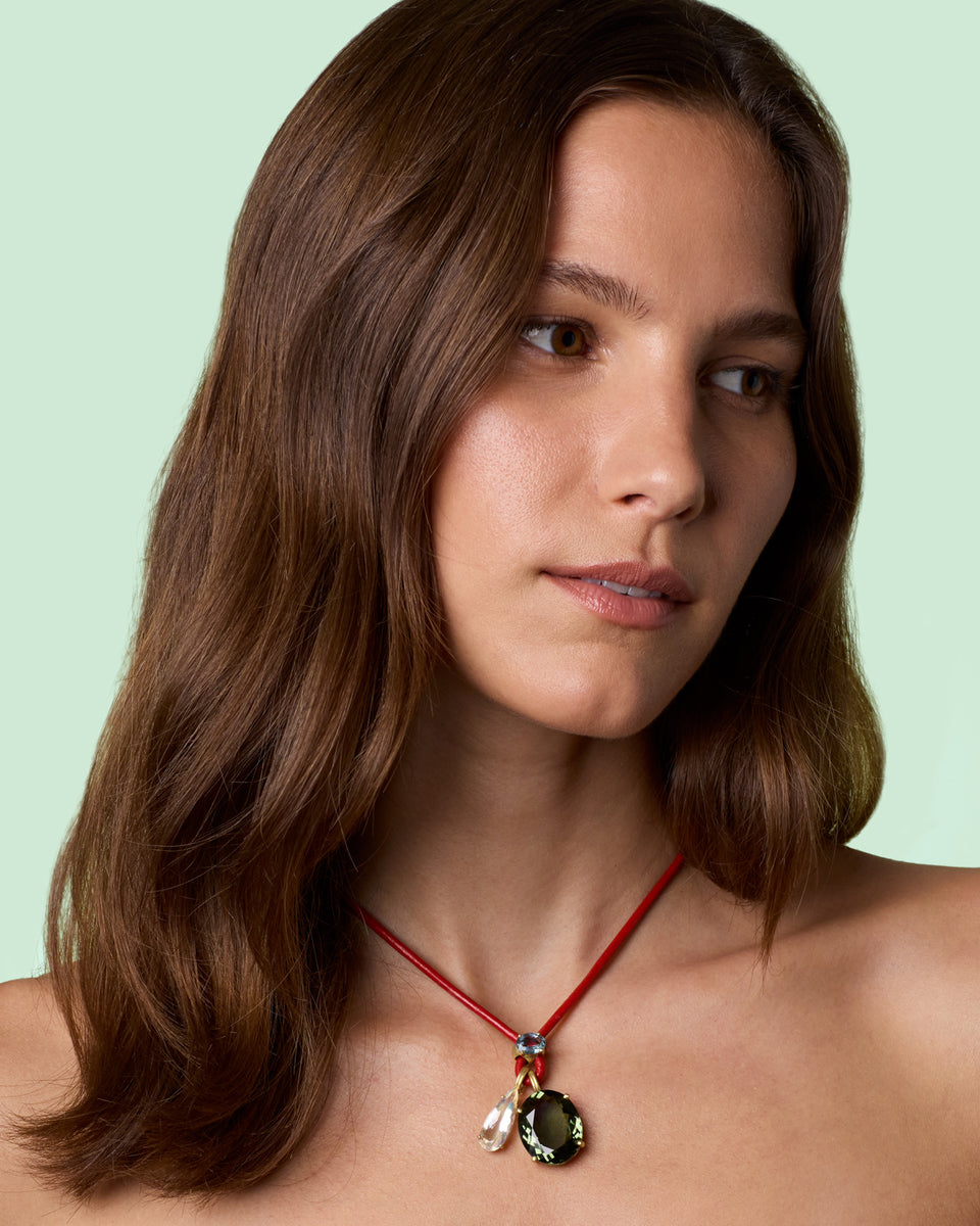 A woman with shoulder-length brown hair wearing a red cord necklace with a dark green oval pendant and small moonstone, set against a solid pale mint green background.
