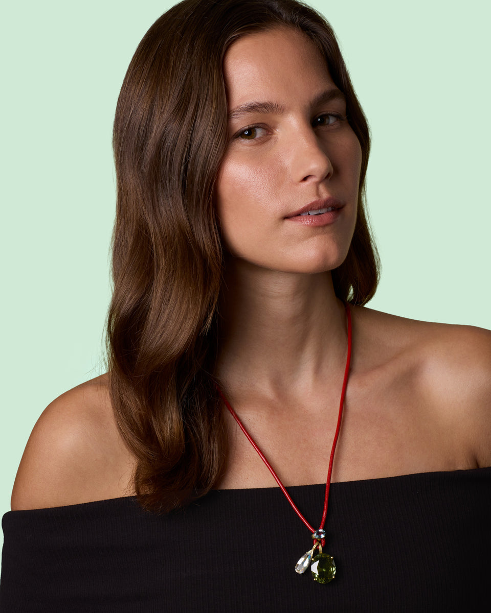 A woman with long brown hair models a red cord necklace with a green and moonstone pendant , set against a solid pale mint green background.