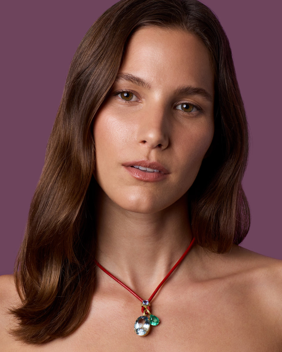 A woman with shoulder-length brown hair wears a red cord necklace with a pendant featuring green and iridescent blue-green stones, set against a solid mauve-purple background.