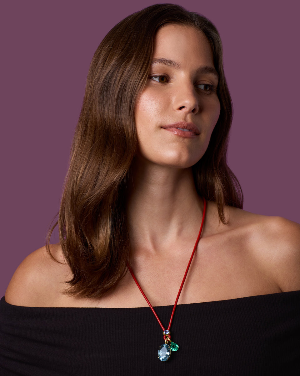 A woman with shoulder-length brown hair models a red cord necklace featuring a pendant with light blue and green faceted gemstones. She is wearing a black off-the-shoulder top against a solid dark purple background.