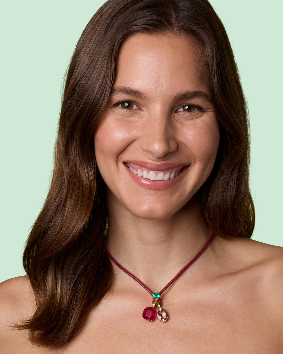 A smiling woman with long brown hair models a cherry-themed necklace featuring a dark red cord, a green top bead, and two dangling pink and red charms, one red and one pink, against a solid pale mint green background.