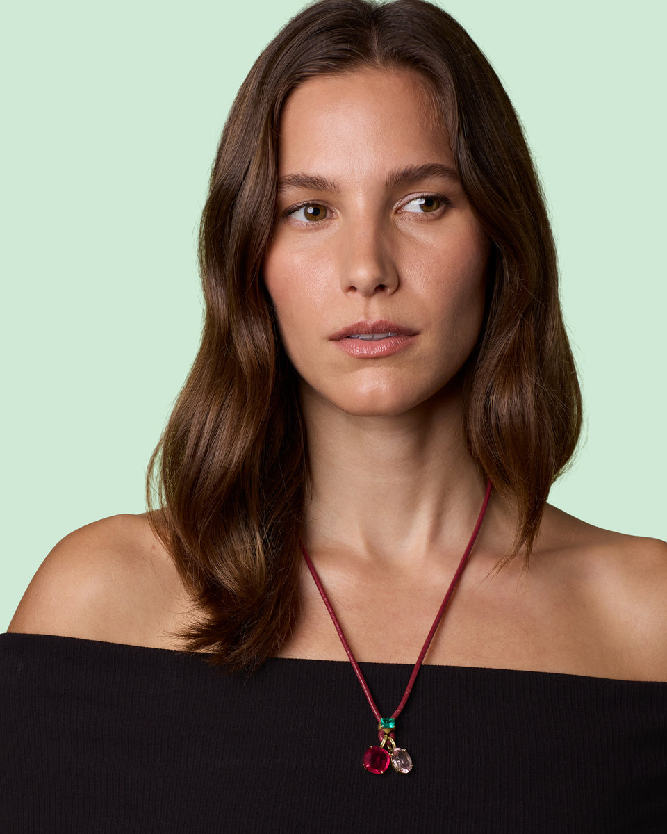 A woman with long brown hair wearing a red cord necklace with a red and pink pendant, shown from the chest up against a pale mint green background.