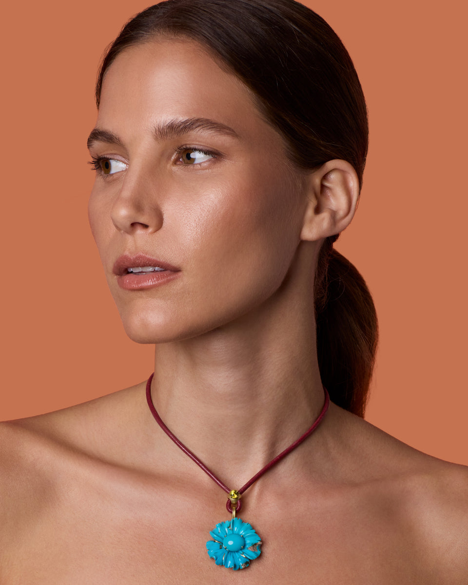 A woman with dark hair tied back wears a turquoise flower-shaped pendant necklace on a dark red cord, set against a solid terracotta background.