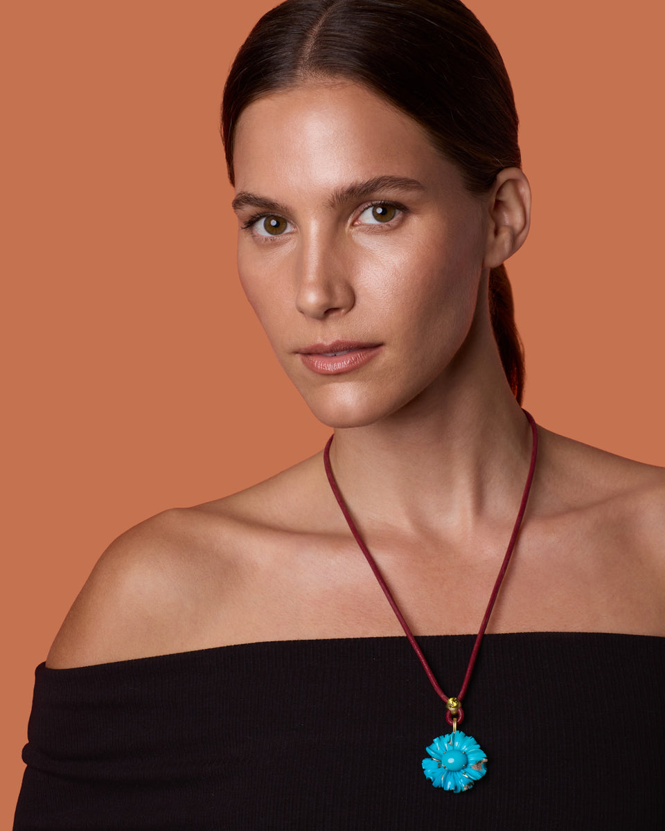 A woman with dark hair pulled back wears a red cord necklace with a bright turquoise flower-shaped pendant, set against a solid warm orange background.