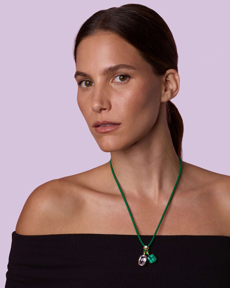 A woman with dark hair tied back wears a green cord necklace with a small green and silver pendant, photographed against a solid light purple background.