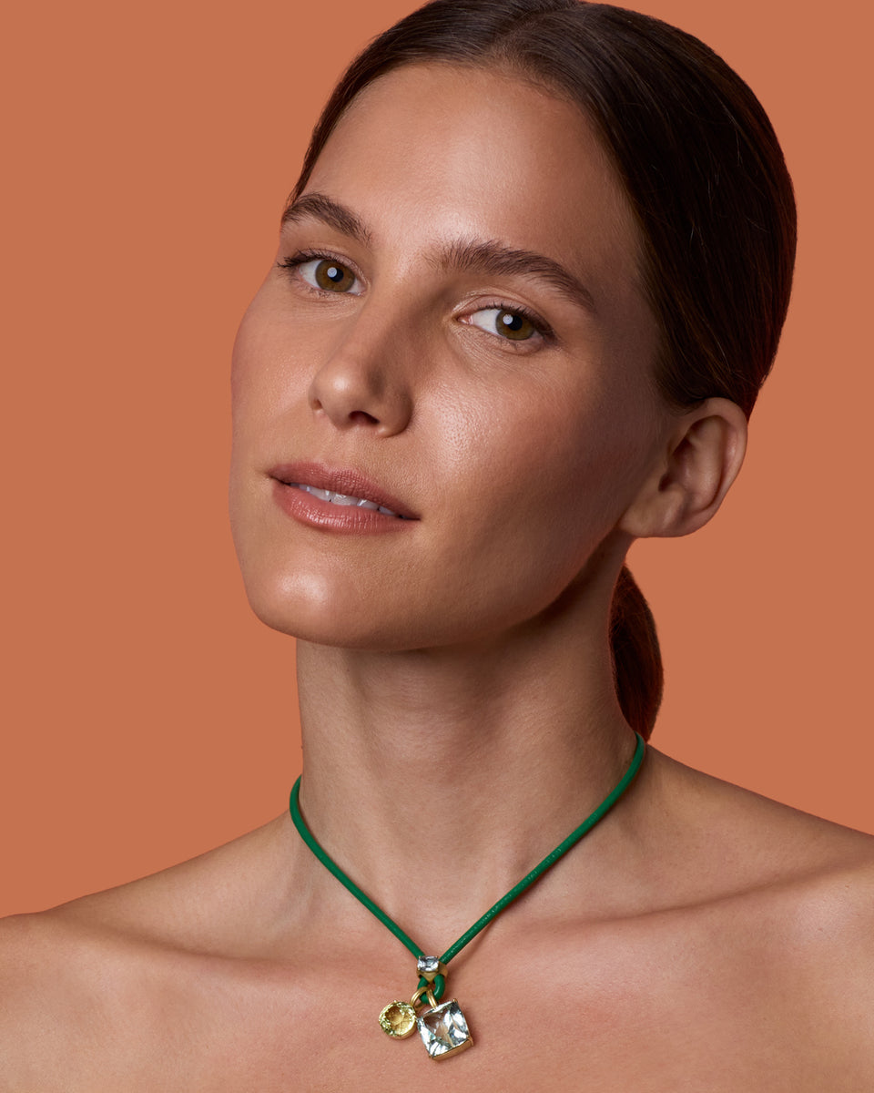 A close-up of a woman with dark hair tied back, wearing a green cord necklace with a small gold and clear sapphire pendant, set against a solid warm orange background.