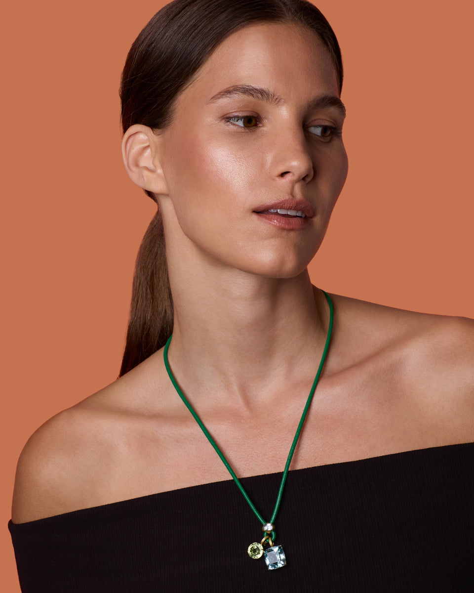 A woman with her hair in a ponytail models a green cord necklace with a small charm pendant against a solid terracotta background.