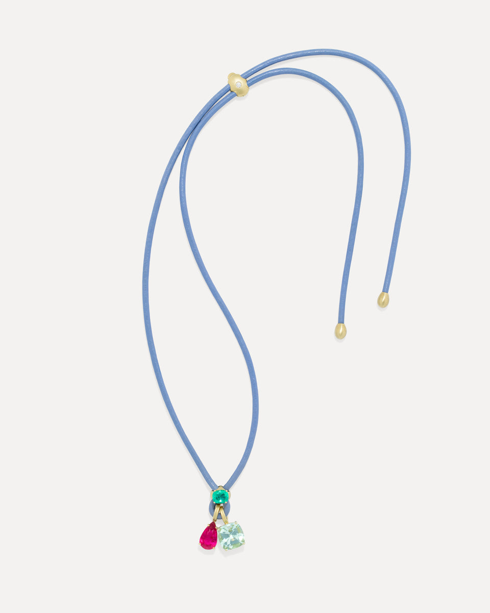 A double-strand blue cord necklace with gold-toned hardware and a small dangling charm featuring green, pink, and light blue heart-shaped stones, shown on a light gray background.