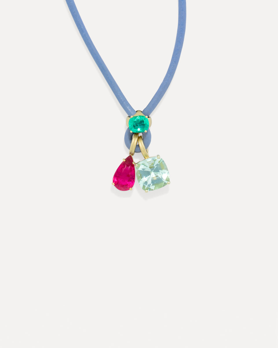 A delicate blue cord necklace featuring a gold-tone pendant with a small green gemstone and two dangling teardrop-shaped gemstones in pink and light green, set against a plain off-white background.