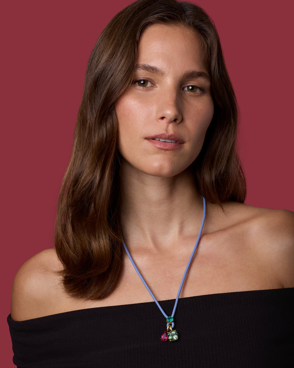 A woman with shoulder-length brown hair models a purple cord necklace with a small colorful pendant against a solid dark red background.