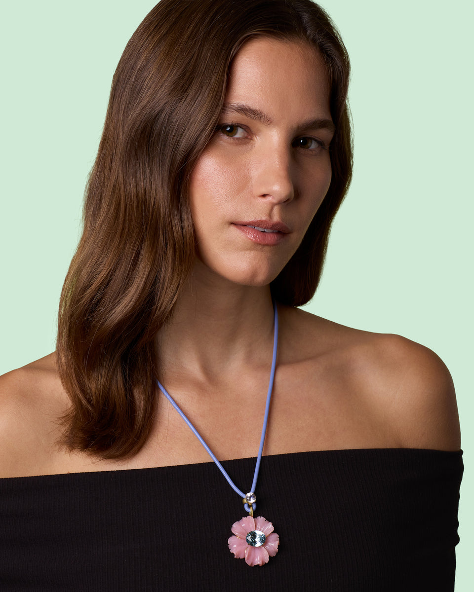 A woman with brown hair wearing a pink flower pendant necklace on a light purple cord, styled with a black off-the-shoulder top against a pale mint green background.