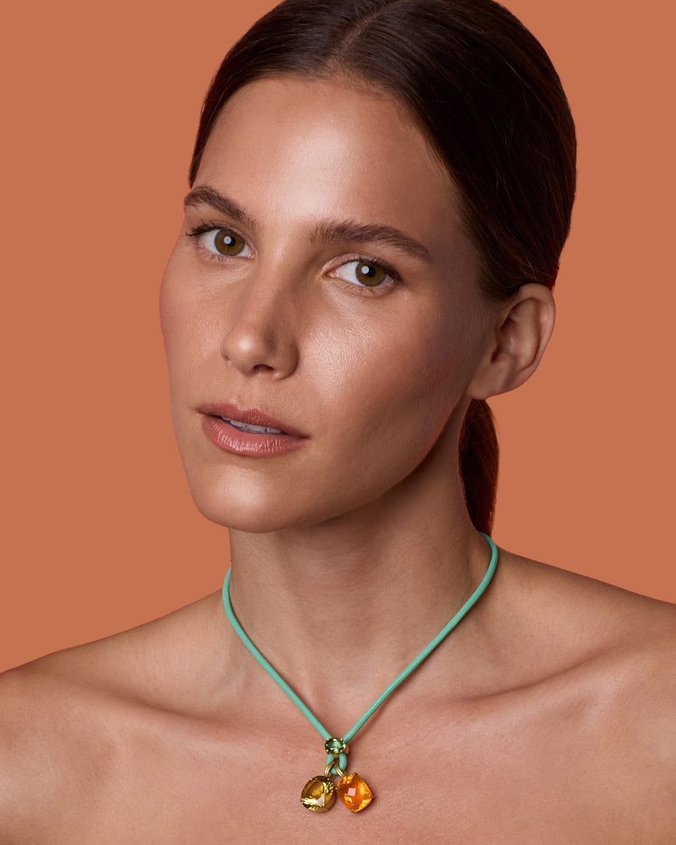 A close-up portrait of a woman wearing a mint green cord necklace with two yellow and orange colored pendants, set against a solid terracotta background.