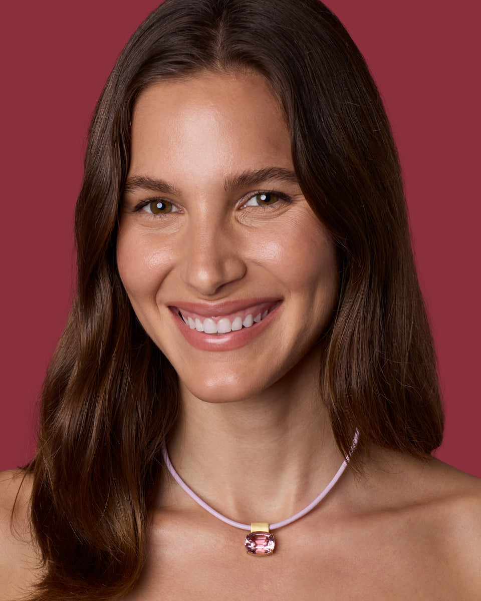 A smiling woman with long brown hair wears a delicate light pink cord necklace with a gold-toned clasp and a pink gemstone pendant, set against a solid dark pink background.