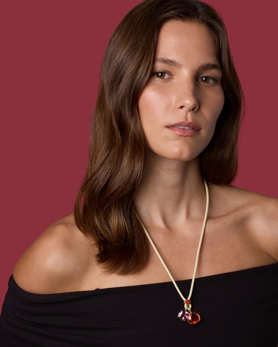 A close-up portrait of a woman with brown hair wearing a delicate gold cord necklace with a small red and amber  pendant, set against a solid dark red background.