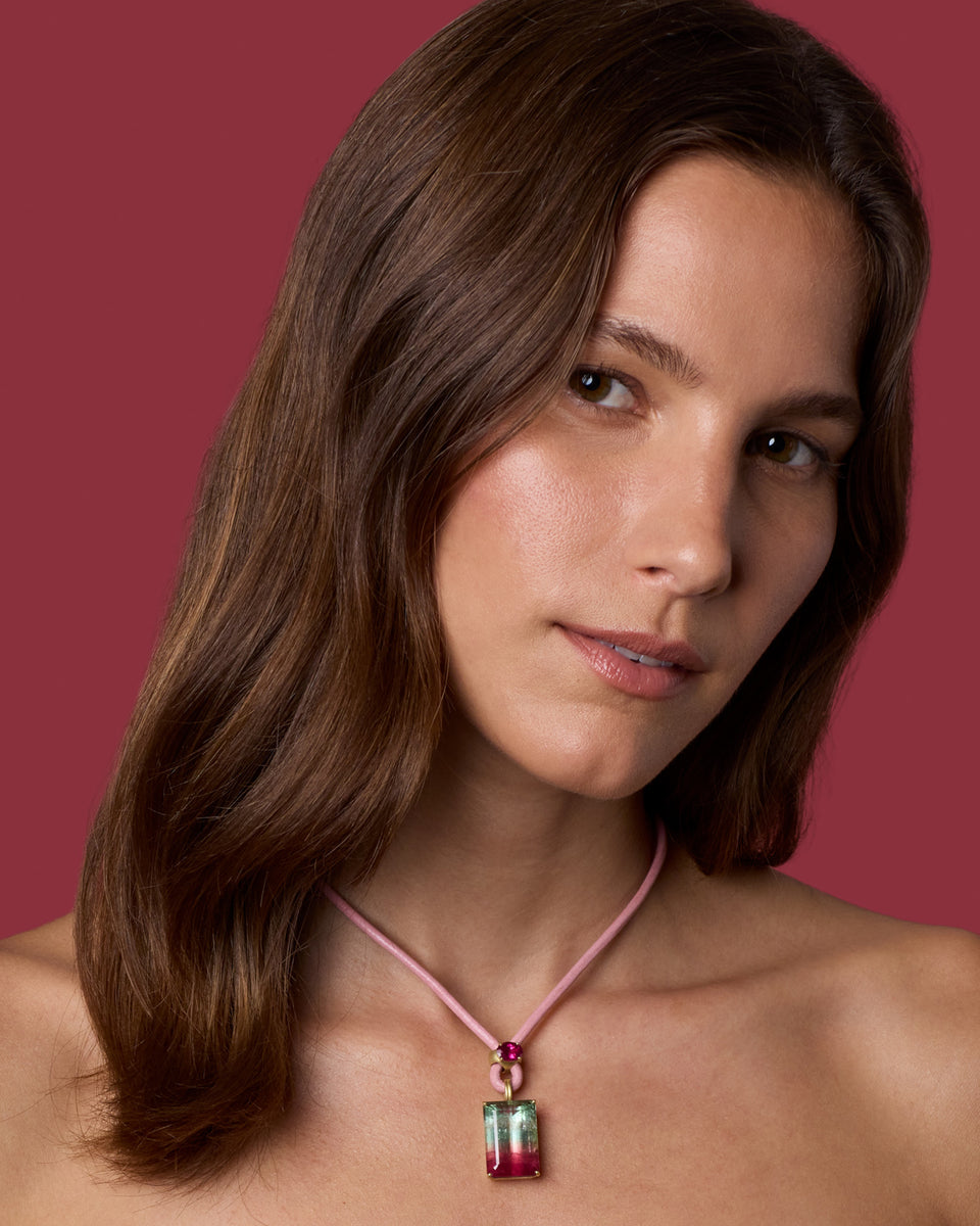 A woman with long brown hair wears a pink cord necklace with a rectangular pendant featuring green, red, and pink gradient facets, set against a solid dark pink background.