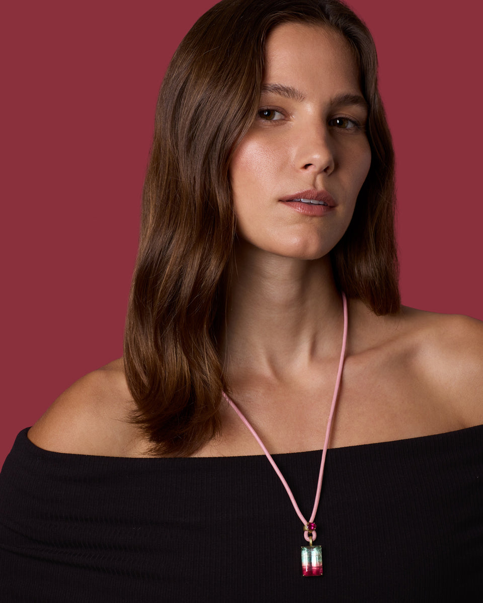 A woman with long brown hair models a pink cord necklace with a small pendant featuring red, white, and green details, set against a solid dark red background.