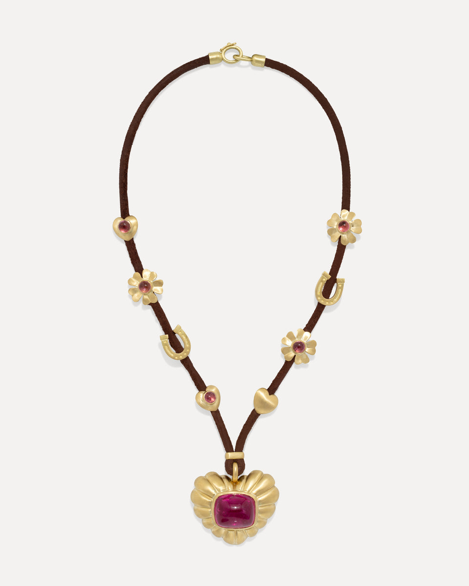 A gold and brown necklace featuring a central gold shell pendant with a magenta cabochon stone, accented by small gold horseshoes, flower motifs, and heart charms on a dark brown cord, set against a light gray background.