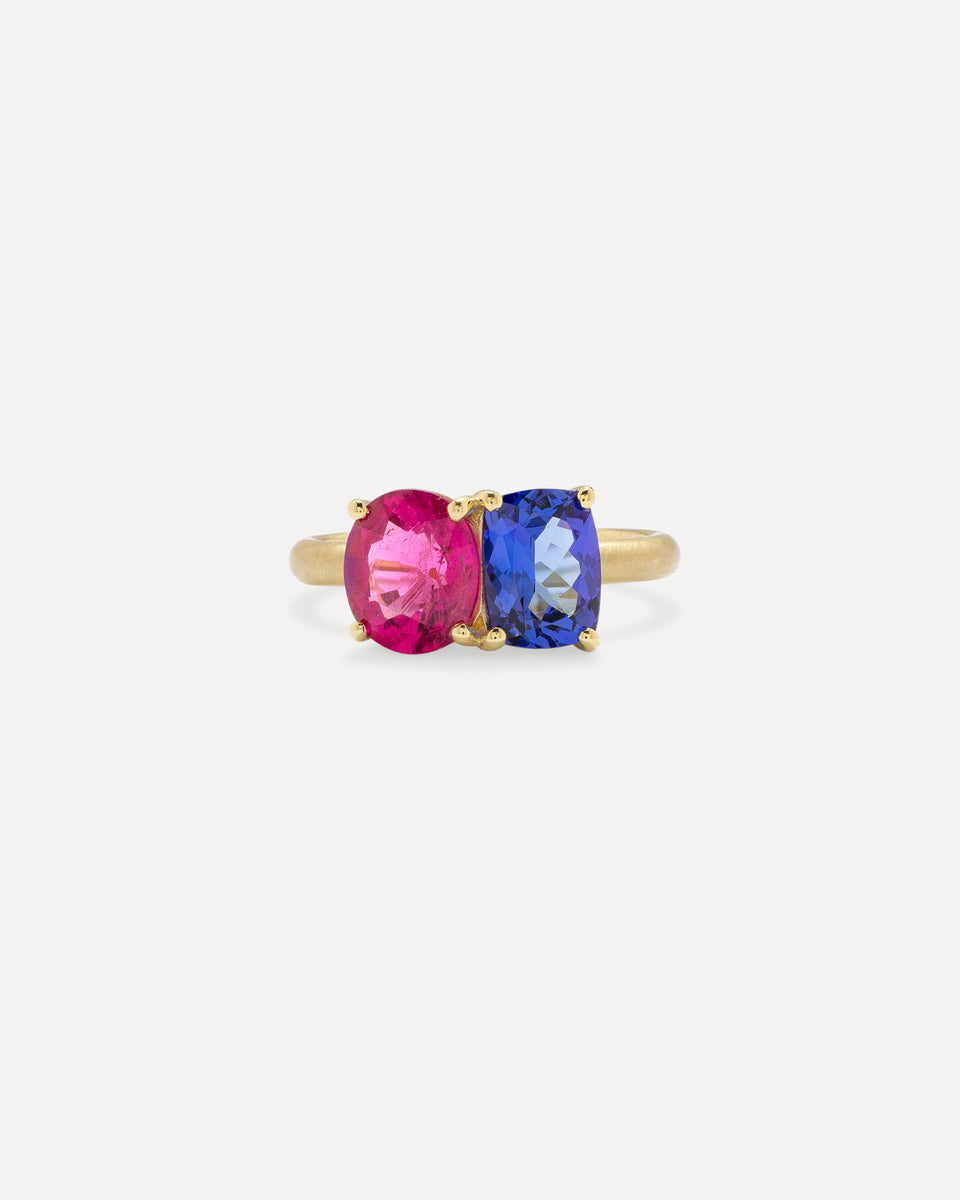 A gold ring featuring two oval-cut gemstones, one vibrant pink and one deep blue, set against a clean off-white background.