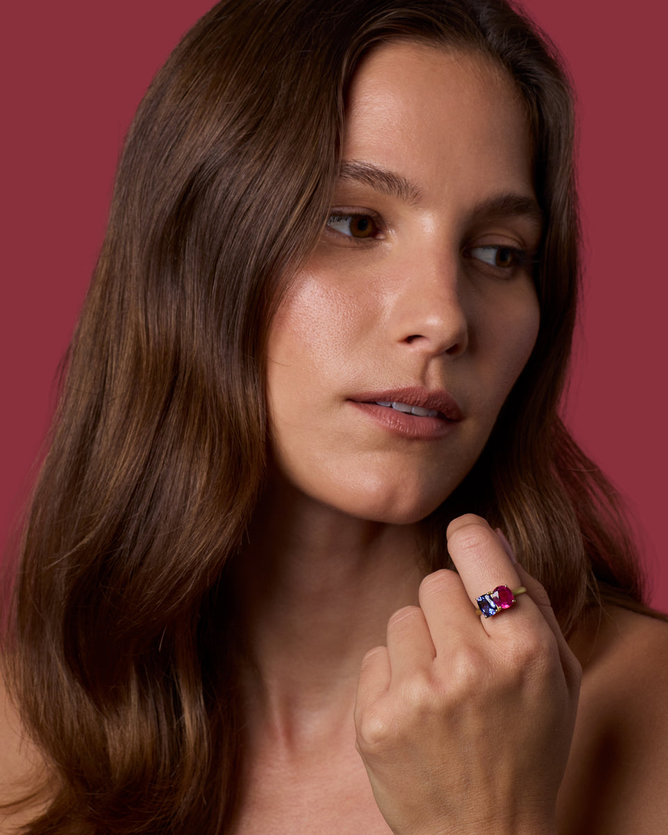 A close-up portrait of a woman with long brown hair wearing a ring with a red and blue gemstone on a pinkish-red background.